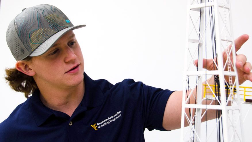 Photo of Nick Lapinski in a baseball cap demonstrating with a model oil rig