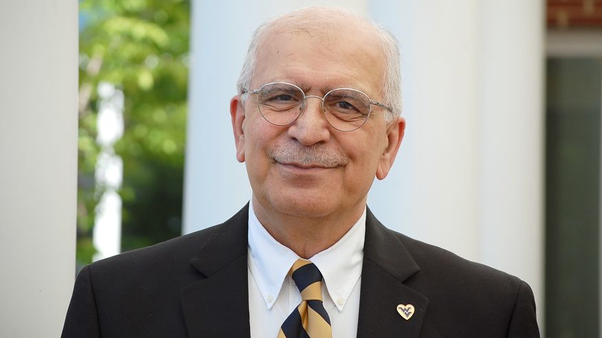 An image of Sam Ameri in a black suit wearing a gold and blue stripped tie, a white dress shirt, and a wvu heart pin on his breast.