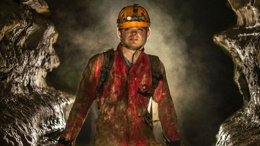 Ryan Maurer wears orange overalls and a yellow hard had with a head lamp, standing inside of a cave with two large rock walls on either side of him