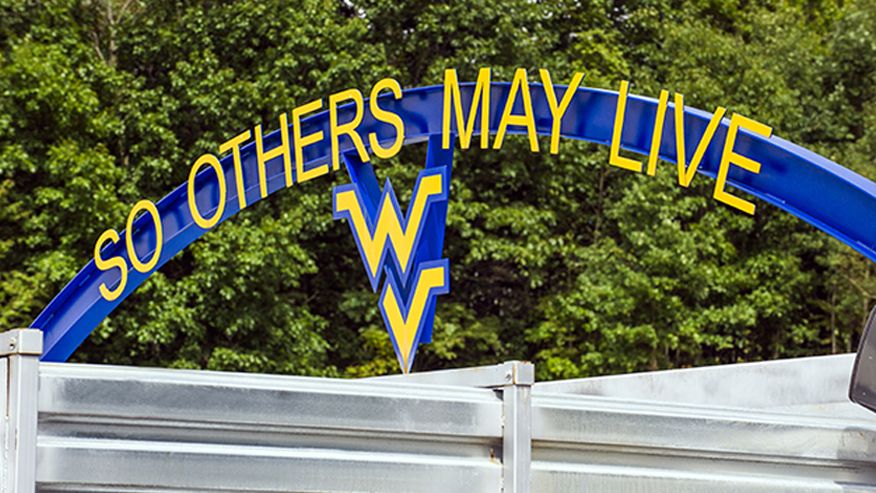 An image of the Sago Mine entrance with a blue WVU sign above it with the words "So others may live" and a flying WV logo in gold