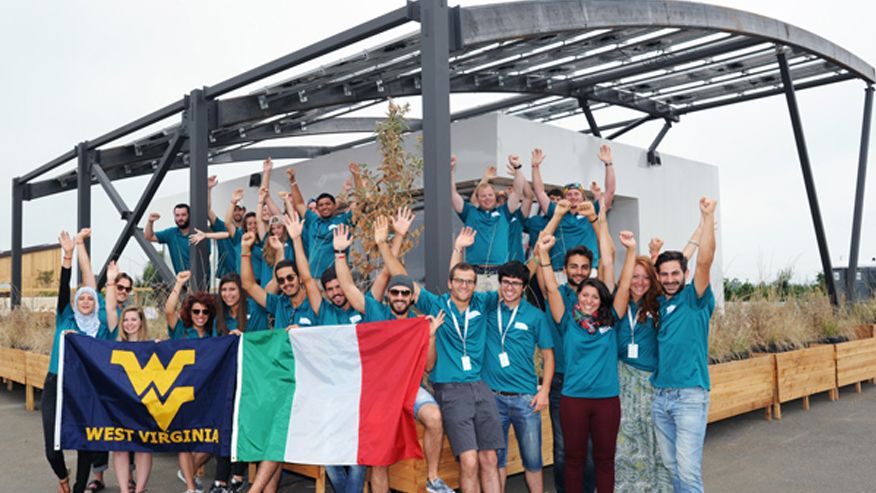 A photo of the University Tor Vergata and West Virginia University 2015 Solar Decathlon team standing in front of their fully built Solar house and waving both the WVU Flag and Italian Flag