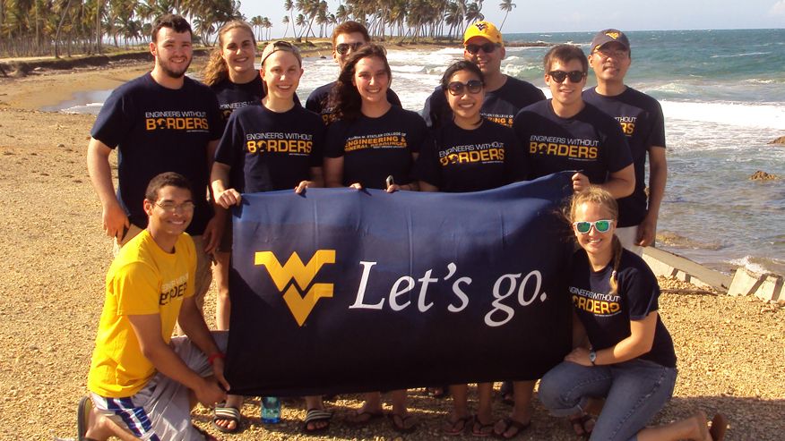 The Engineers without Borders Group on a beach together holding up the a blue flag with a gold WVU logo and the words Let's Go on it